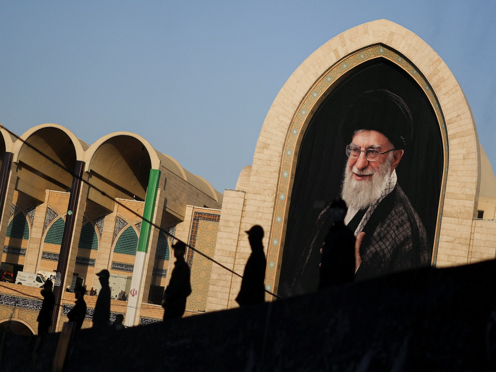 People stand silhouetted against a large depiction of the late Supreme Leader Ayatollah Ali Khamenei in the background during a public farewell ceremony to the late Supreme Leader (Photo/Reuters)